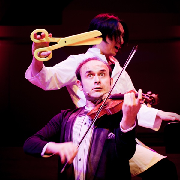 Joo holding a pair of bright yellow oversize scissors over the head of Igudesman, who is playing the violin.