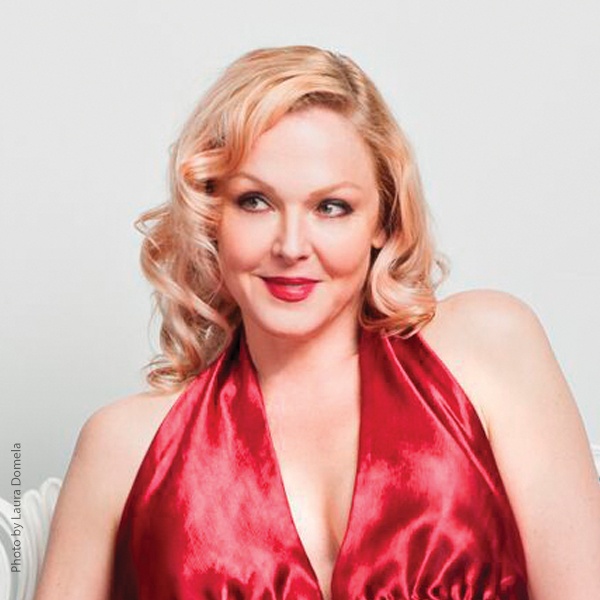 Storm Large in a red dress sitting on a chair and smiling.