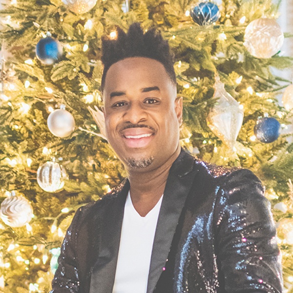 Damien Sneed in a dark jacket smiling in front of a brightly lit and decorated Christmas tree.