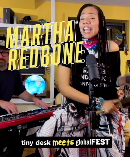 Martha Redbone singing from her home studio. Behind her sits her keyboardist and guitarist, the walls a warm yellow and an illuminated globe in the background.